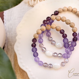 Könnte beinhalten: Drei Armbänder mit lila und weißen Perlen. Die Armbänder sind aus Holz, Glas und undurchsichtigen lila Perlen gefertigt. Die Armbänder sind übereinander gestapelt.