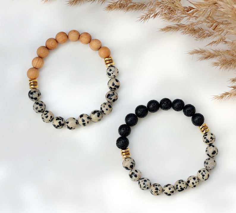 May include: Two bracelets made with natural stones. One bracelet is made with brown wood beads and white and black dalmatian jasper beads. The other bracelet is made with black lava beads and white and black dalmatian jasper beads. Both bracelets have gold spacers.
