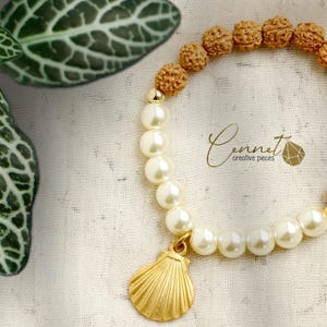 Könnte beinhalten: Ein Armband mit abwechselnden Abschnitten aus hellbraunen, strukturierten Perlen und cremefarbenen Perlen. Ein goldfarbener Muschelanhänger hängt am Perlenabschnitt. Das Armband liegt vor einem weißen Stoffhintergrund. Der Text "Cennet creative pieces" ist ebenfalls sichtbar.