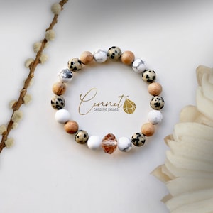 Könnte beinhalten: Ein Armband mit einer Mischung aus Natursteinperlen in Weiß, Braun und gesprenkelten Mustern. Das Armband hat eine zentrale facettierte Perle. Das Bild enthält auch einen Zweig mit Kätzchenweide und den Text "Cennet creative pieces."