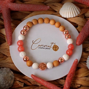 Könnte beinhalten: Ein Armband mit Holzperlen, weißen Lavasteinen, roségoldenen Perlen und rosafarbenen Steinperlen. Das Armband liegt auf einem weißen Hintergrund mit dem Text "Cennet Creative Pieces".