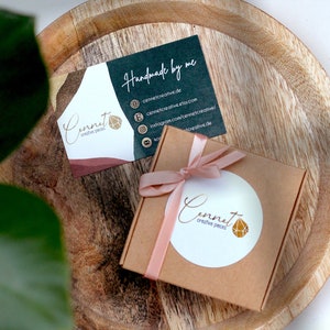 May include: A small, brown gift box with a pink ribbon and a white circle sticker with the text "Cennet creative pieces" and a gold geometric design. The box is sitting on a wooden surface next to a business card with the text "Handmade by me" and the company name "Cennet creative pieces".