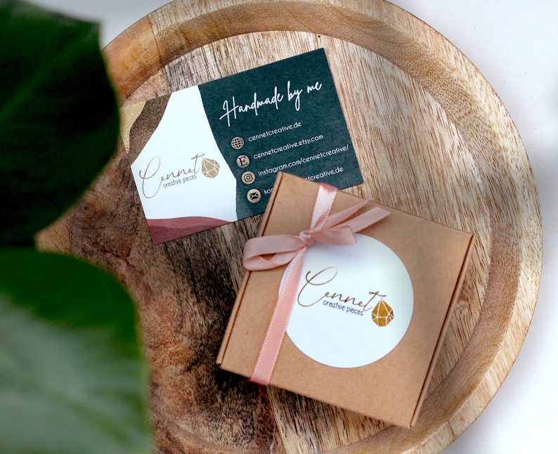 May include: A small, brown gift box with a pink ribbon and a white circle sticker with the text "Cennet creative pieces" and a gold geometric design. The box is sitting on a wooden surface next to a business card with the text "Handmade by me" and the company name "Cennet creative pieces".