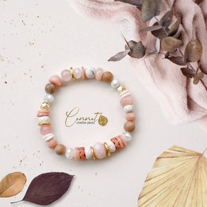 May include: A pink, white and gold beaded bracelet with a wooden bead and a gold charm. The bracelet is on a white background with a piece of fabric in the background.