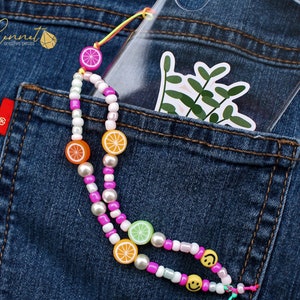 Puede incluir: Una funda de teléfono transparente con una pegatina de diseño de hojas verdes en el bolsillo trasero de unos vaqueros azules. Una correa de teléfono con cuentas rosa y blanca con abalorios de frutas de colores cuelga de la funda.
