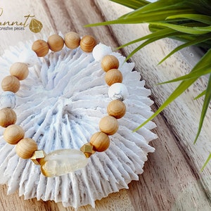 Könnte beinhalten: Ein Armband aus Holz- und weißen Perlen mit einem goldfarbenen, facettierten, klaren Quarz-Anhänger. Das Armband liegt auf einer weißen korallenartigen Oberfläche.