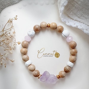Peut inclure: Un bracelet composé de perles de bois naturel, de pierres blanches et roses et d'un grand charme en cristal d'améthyste. Le bracelet est sur une surface blanche avec un petit bouquet de fleurs séchées.