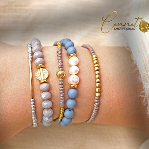 Könnte beinhalten: Ein Set aus fünf Armbändern mit verschiedenen Materialien und Farben. Die Armbänder umfassen eine silberne Kette, ein Armband mit grauen und weißen Perlen, ein Armband mit grauen Perlen, ein Armband mit hellblauen Perlen und ein Armband mit goldenen Perlen.