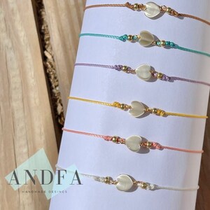 Puede incluir: Un conjunto de diez pulseras con cordones de diferentes colores y un dije de corazón blanco en cada una. Las pulseras están dispuestas en fila sobre una superficie de madera.