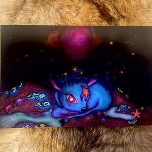 May include: A blue and purple illustration of a deer with a star on its forehead, sleeping on a bed of stars and flowers. The deer has a pink eye and a pink nose. The background is a dark blue with a pink planet.