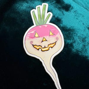 May include: A Halloween-themed sticker featuring a smiling turnip with a jack-o'-lantern face. The turnip has a pink top with green stems and a cream-colored body. The sticker is on a dark green velvet surface.