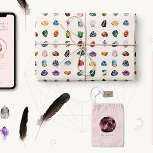 May include: A flat lay image of a pink and white gift bag with a crystal pattern, a pink and white card with the text "Healing Crystals for Emotional Balance", a pink and white phone case with the text "A Beginner's Guide to Healing Crystals", a pink and white pouch with the text "la moon", and a black feather.