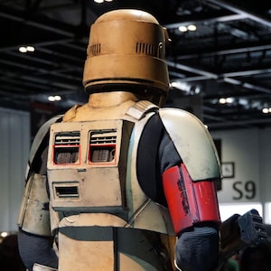 Puede incluir: Una figura de acción de Star Wars de color beige y marrón con un casco y un brazo rojo y negro. La figura tiene una mochila con dos compartimentos rectangulares en la espalda.