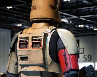 Könnte beinhalten: Eine beige und braune Star Wars Actionfigur mit Helm und einem rot-schwarzen Arm. Die Figur hat einen Rucksack mit zwei rechteckigen Fächern auf dem Rücken.