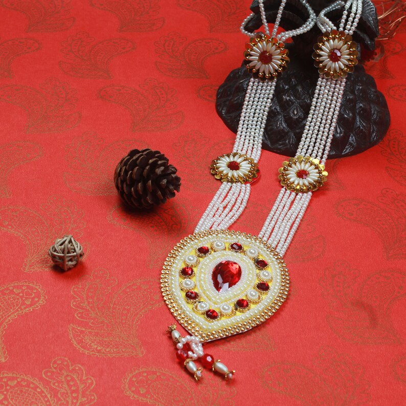 Op de afbeelding: Een witte parelketting met rode en gouden accenten. De ketting heeft een grote, sierlijke hanger met een rode edelsteen in het midden. De ketting wordt gepresenteerd op een rode achtergrond.