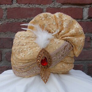 Op de afbeelding: Een gouden turban met een witte veer en een rode juweelaccent. De turban is versierd met gouden borduurwerk en kralen.