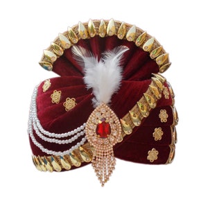 Op de afbeelding: Een maronkleurige fluwelen turban met gouden versieringen en een parelketting. Een grote broche met een rode steen is aan de turban bevestigd.