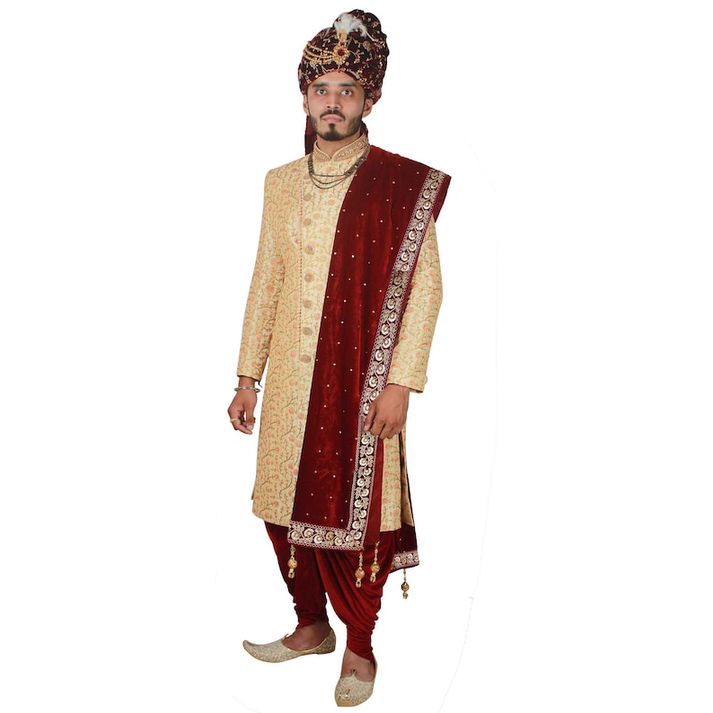 May include: A man wearing a gold embroidered sherwani with a maroon velvet shawl and a gold turban. The shawl has gold embroidery and tassels.