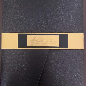 May include: A black, textured envelope with a gold band wrapped around it. The gold band has a black rectangle with the text "Watkins Rd. CHURCH OF CHRIST" in gold lettering.