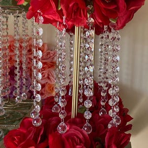 May include: A decorative centerpiece with bright red roses and hanging crystal strands. The arrangement has a gold-coloured stand and a mirrored base, creating a glamorous display. The roses are clustered at the top and base.
