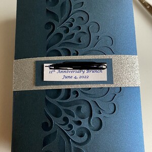 May include: A dark blue invitation with a silver band and a white label that reads "11th Anniversary Brunch June 4, 2022". The invitation has a decorative, laser-cut design.