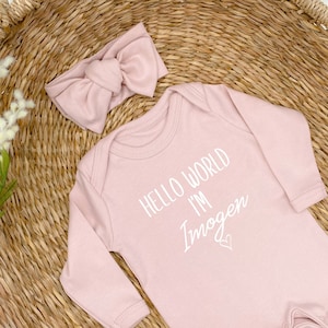 Pode incluir: Um macac&atilde;o rosa para beb&ecirc; com mangas compridas e um gr&aacute;fico branco que diz "Hello World I'm Imogen" com um pequeno cora&ccedil;&atilde;o. Um la&ccedil;o de cabelo rosa est&aacute; inclu&iacute;do.