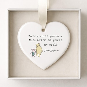 K&ouml;nnte beinhalten: Wei&szlig;es herzf&ouml;rmiges Keramikornament mit cremefarbenem Band. Es zeigt eine Winnie-the-Pooh-Illustration und den Text: &bdquo;To the world you're a Mum, but to me you're my world. Love Josie x.&ldquo;