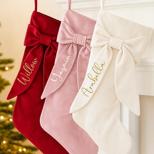 May include: Three velvet Christmas stockings in red, pink, and cream, each with a large bow and a name embroidered in gold: Willow, Yasmin, and Arabella. The stockings hang from a white mantelpiece.