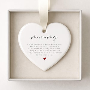 May include: A heart-shaped ceramic ornament, white with a cream ribbon. The word "mummy" is written in cursive. The text reads, "I'm snuggled up warm..." A small red heart is at the bottom. The ornament is presented in a white box.