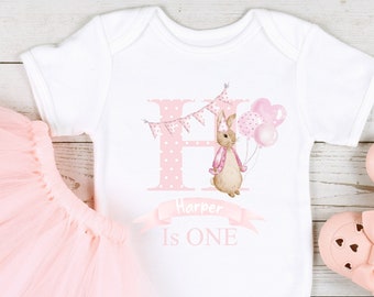 May include: A white onesie with a pink polka dot letter H, a bunny holding balloons, and the text "Harper is ONE". The onesie is paired with a pink tulle tutu and pink baby shoes.