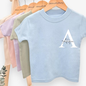 May include: A baby blue t-shirt with the name "Alfie" printed in black script below a large white letter "A". The shirt is one of several pastel-colored t-shirts hanging on wooden hangers, with the brand name visible.