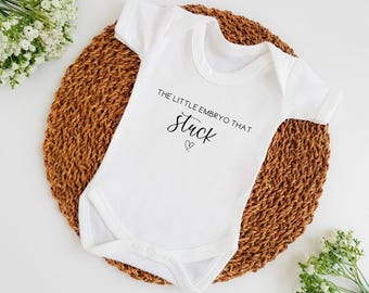 Puede incluir: Un body blanco para bebé con mangas cortas y un gráfico negro que dice "El pequeño embrión que se quedó" con un pequeño símbolo de corazón.