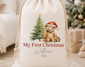 May include: A cream-colored canvas drawstring gift bag featuring a watercolor illustration of a teddy bear wearing a red Santa hat next to a decorated Christmas tree and presents. The text reads My First Christmas, William, and 2026.