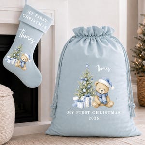 May include: A light blue Christmas stocking and a matching drawstring gift sack. Both items feature a printed illustration of a teddy bear wearing a blue hat and scarf, a decorated Christmas tree, and the name Thomas with My First Christmas text.