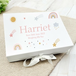 May include: White gift box featuring a pink and gold rainbow, stars, and shooting stars design. The box is personalised with the name "Harriet", the date "Born 26th June 2023", and the weight "Weighing 8lb 9oz".