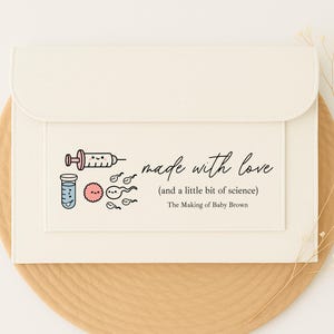 May include: Off-white envelope with a flap, decorated with a syringe, test tube, and sperm cell illustrations. The text says "made with love (and a little bit of science)" and "The Making of Baby Brown."