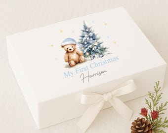 May include: A white gift box with a cream-colored bow. The box is decorated with a watercolor illustration of a teddy bear wearing a blue hat, a Christmas tree, and the text "My First Christmas" and a name. A pine cone and greenery are next to the box.