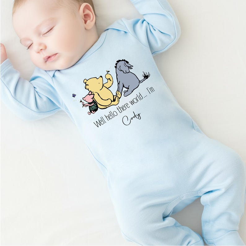 May include: A light blue baby onesie with a Winnie the Pooh, Piglet, and Eeyore graphic. The onesie has the text "Well hello there world... I'm Cody" printed on it.