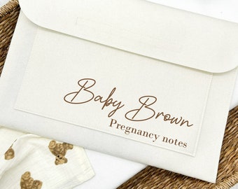 Op de afbeelding: Een witte viltmap met de tekst "Baby Brown Pregnancy notes" in bruine letters. De map ligt in een geweven mand met een witte stof met bruine teddybeer-ontwerpen.