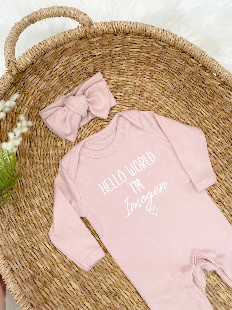 Pode incluir: Um macac&atilde;o rosa para beb&ecirc; com mangas compridas e um gr&aacute;fico branco que diz "Hello World I'm Imogen" com um pequeno cora&ccedil;&atilde;o. Um la&ccedil;o de cabelo rosa est&aacute; inclu&iacute;do.