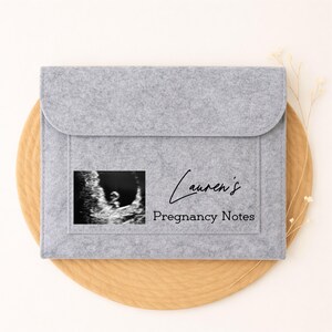 May include: A light grey felt pregnancy notes holder with a flap closure. The holder features a sonogram image and the text "Lauren's Pregnancy Notes" in black script. The holder is on a round, light brown surface.