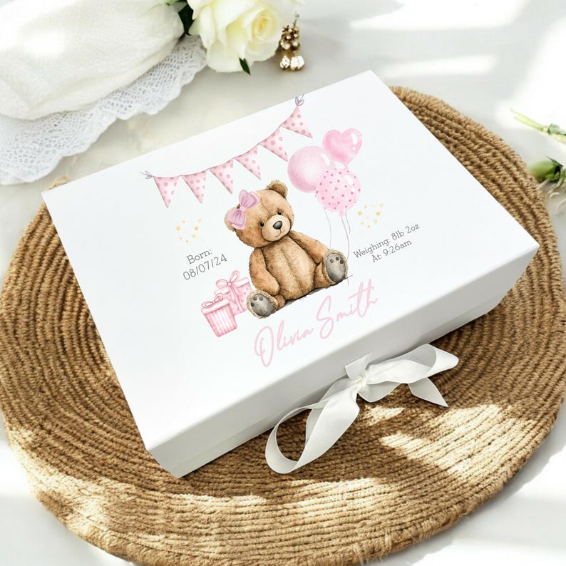 May include: A white keepsake box with a teddy bear illustration, pink balloons, and a banner. The box is decorated with the text "Born 08/07/24", "Weighing 8lb 2oz At 9:26am", and the name "Olivia Smith". A white ribbon is tied around the box.