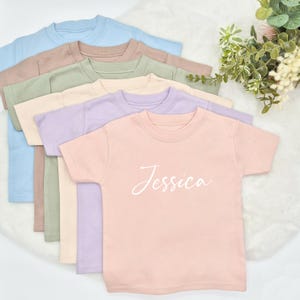 May include: A stack of six plain t-shirts in various pastel colours, including light blue, beige, green, cream, lavender, and pink. The pink t-shirt has the name "Jessica" written in white cursive script.