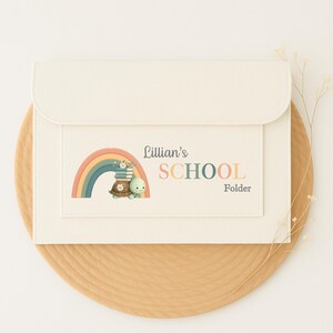 May include: Cream-coloured school folder with a flap closure. The front features a rainbow, a turtle with books, and the text "Lillian's SCHOOL Folder" in various colours. The folder is on a woven, round, tan placemat.