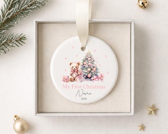 Könnte beinhalten: Runder, weißer Keramik-Weihnachtsschmuck mit rosa Design. Der Schmuck zeigt einen Teddybären, einen Weihnachtsbaum und Geschenke. Der Text auf dem Schmuck lautet "My First Christmas" und "Name" mit dem Jahr 2026.