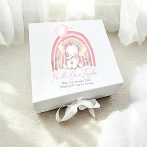 May include: White gift box with a pink and white rainbow design featuring a small elephant holding a pink balloon. The box is tied with a white ribbon and has the text "Millie Mia Taylor" and "Born: 3rd January 2024 Weighing 7lb 3oz At 9:23am" printed on the front.