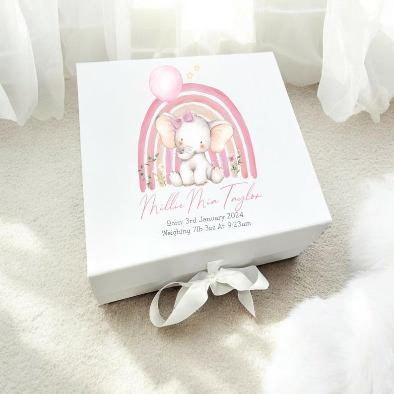 May include: White gift box with a pink and white rainbow design featuring a small elephant holding a pink balloon. The box is tied with a white ribbon and has the text "Millie Mia Taylor" and "Born: 3rd January 2024 Weighing 7lb 3oz At 9:23am" printed on the front.