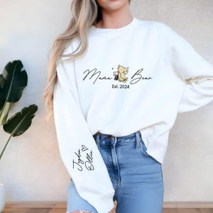 May include: White sweatshirt with the text "Mama Bear Est. 2024" and a cartoon image of Winnie the Pooh and Piglet. The left sleeve has the text "Jayden Olivia" with a heart symbol.