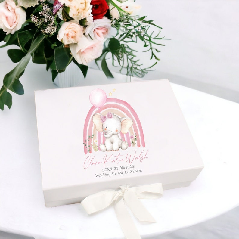 May include: A white gift box with a pink and white rainbow and a cute elephant holding a pink balloon. The box is tied with a white ribbon and has the text "Clara Katie Walsh BORN: 23/08/2023 Weighing 5lb 4oz At 9:26am"