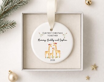 May include: A white ceramic Christmas ornament with a cream-colored ribbon. The ornament features a giraffe family wearing Santa hats, the text "OUR FIRST CHRISTMAS TOGETHER", and the names "Mummy, Daddy and Sophia" with the year 2026.